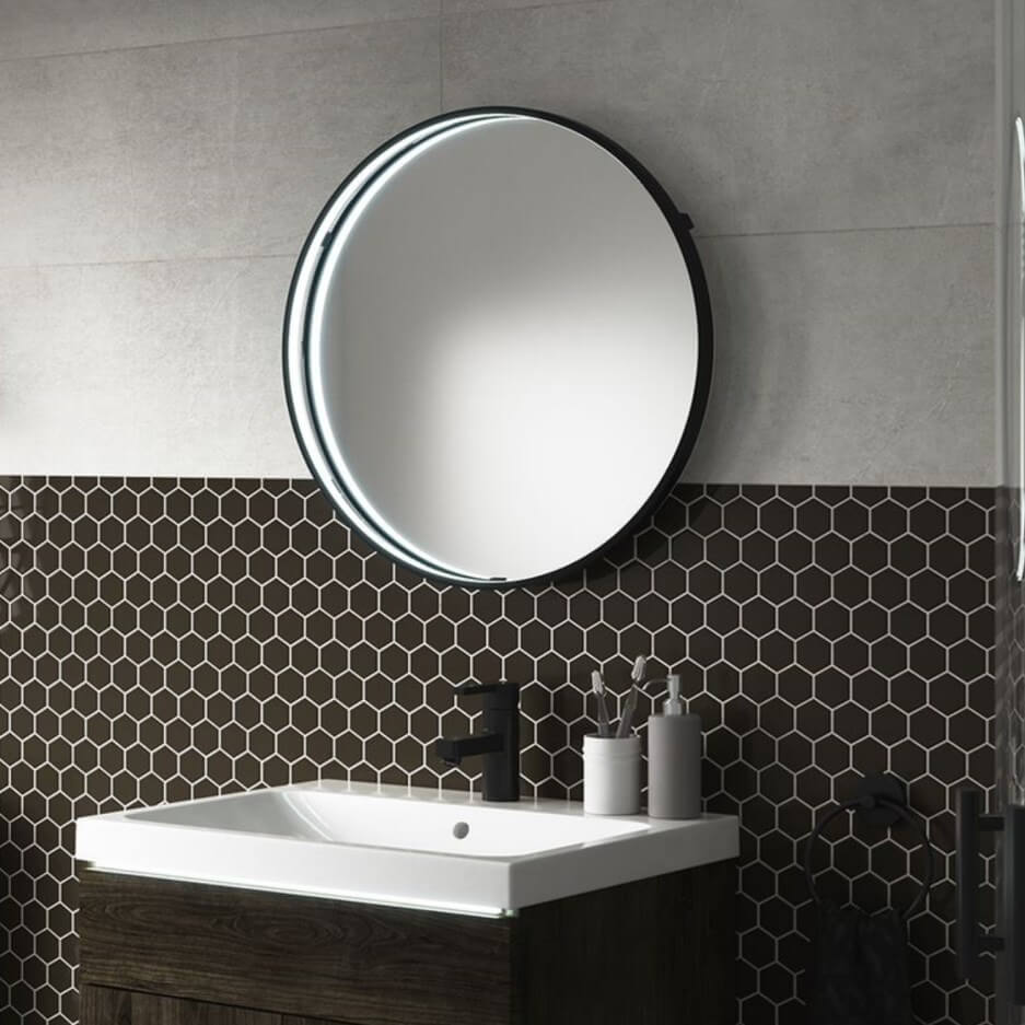 Round, black mirror with light set above a bathroom basin unit, with grey & black tiled wall behind.